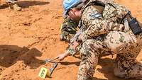 Zwei Soldaten knien im Gelände vor einem Beweisstück mit Nummer und Fotolineal.