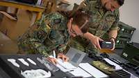 Zwei Soldaten stehen gebückt vor den einzelnen Zeilen eines Gerätes und haben eine Liste auf dem Tisch.