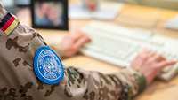 Ein Frau in Flecktarn-Uniform sitzt an einem Schreibtisch und tippt auf einer Tastatur, auf ihrem Arm ein Abzeichen der UN.