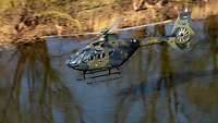 Ein Hubschrauber fliegt über einem Fluss. Bäume spiegeln sich im Wasser.