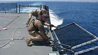 Zwei Soldaten hocken sich an den Rand des Flugdecks und klappen gemeinsam mit Seilen vorsichtig die Seereling ab